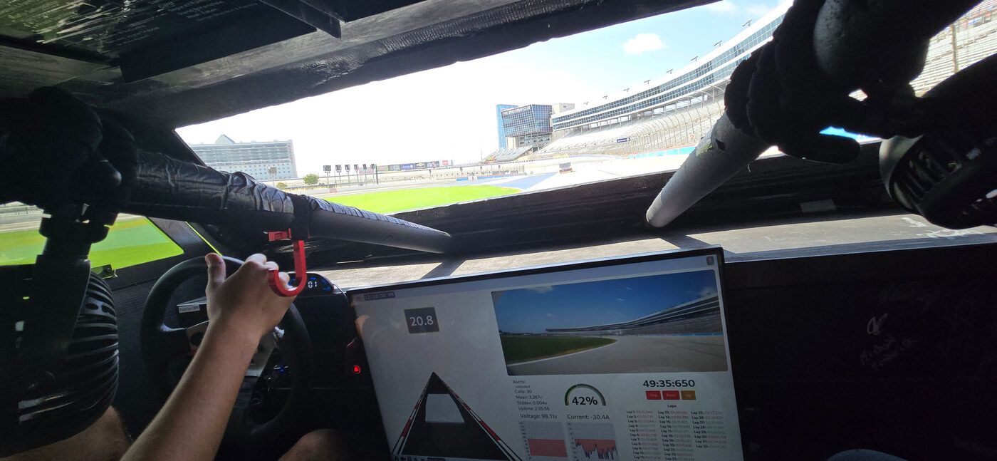 View from inside the solar car on the speedway. A driver's hand rests on the
                    steering wheel; the dashboard screen shows a live track camera, lap times, a
                    42 percent state-of-charge gauge, pack voltage and current, and per-cell
                    battery statistics.